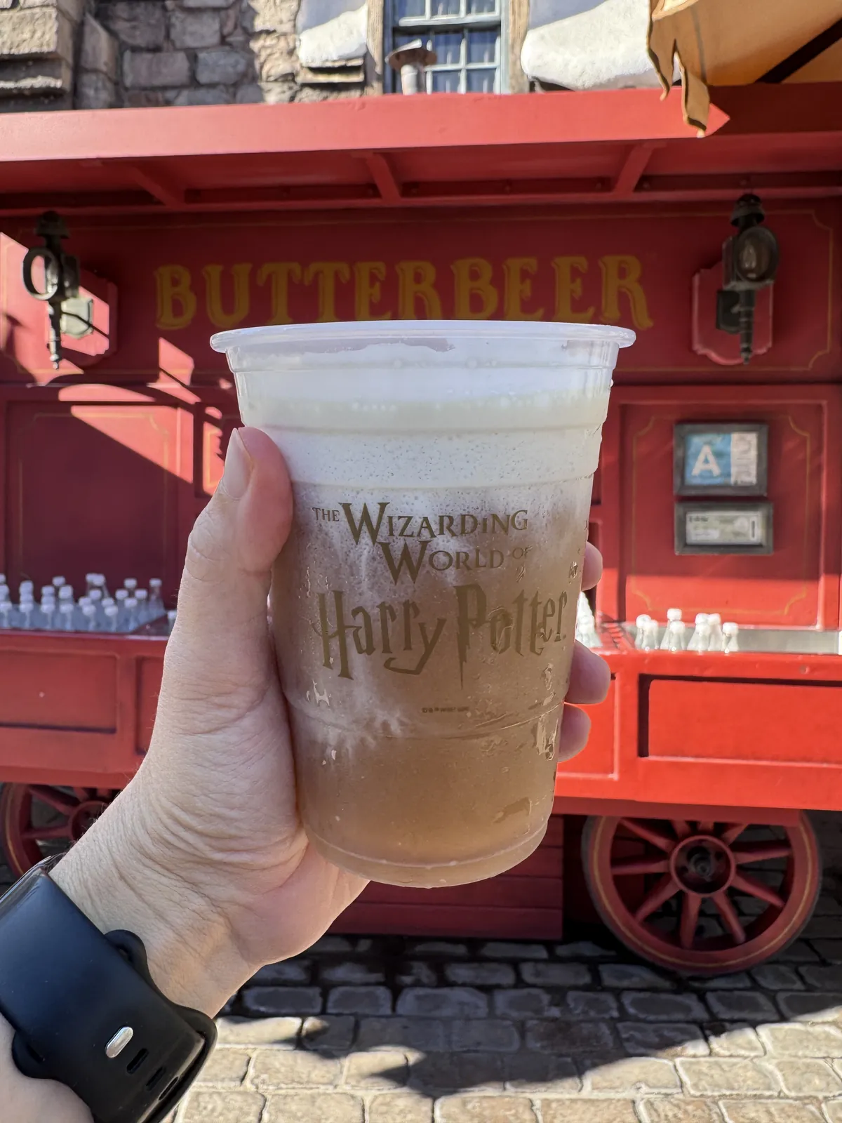Cold Butterbeer in a Wizarding World of Harry Potter cup in front of the red Butterbeer cart in Hogsmeade at Universal Studios Hollywood